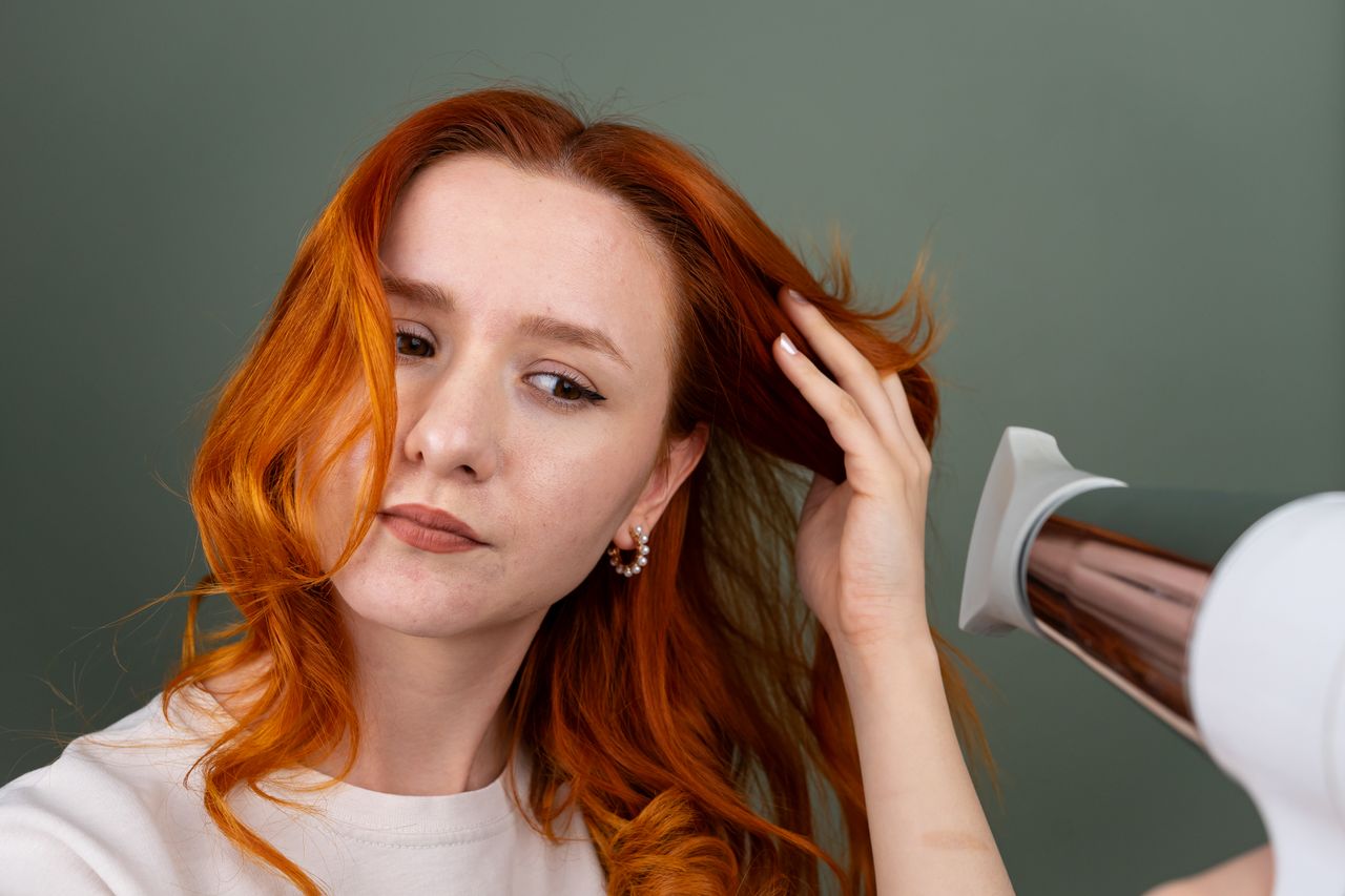 Close-up of a woman using a hairdryer to style her long red hair indoors.